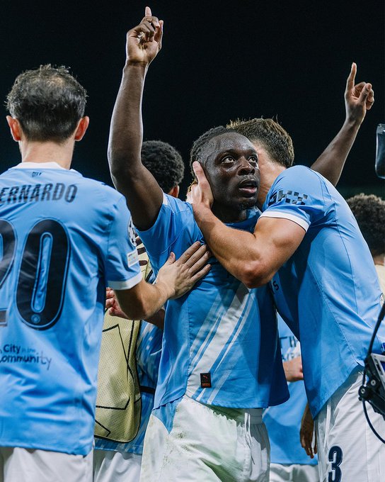 Jeremy Doku wearing a light blue Manchester City jersey, raising both arms in celebration on a soccer field. Other players in similar jerseys, including one with "BERNARDO" and the number 20 on the back, are also celebrating, with some raising their arms and others embracing.