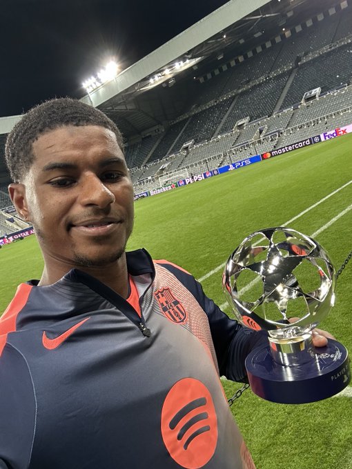 Marcus Rashford standing on a soccer field holding the UEFA Champions League trophy. He is wearing a dark Nike jersey with orange accents and the FC Barcelona logo. The stadium seats and field are visible in the background under stadium lights.