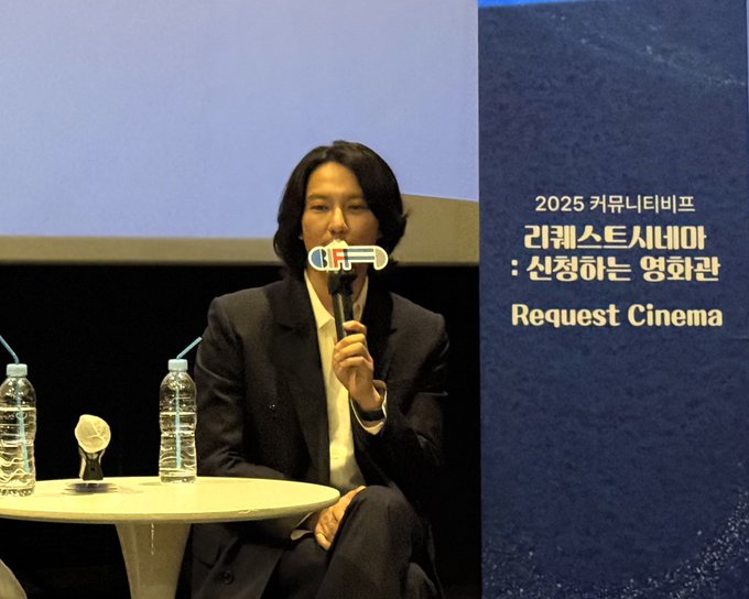 Kim Nam-gil sitting at a table, holding a microphone, wearing a dark suit and white shirt. Two water bottles and a tissue box are on the table. Text overlay reads "2025 부산국제영화제 Request Cinema" and "김래스시네마: 신청하는 영화관".