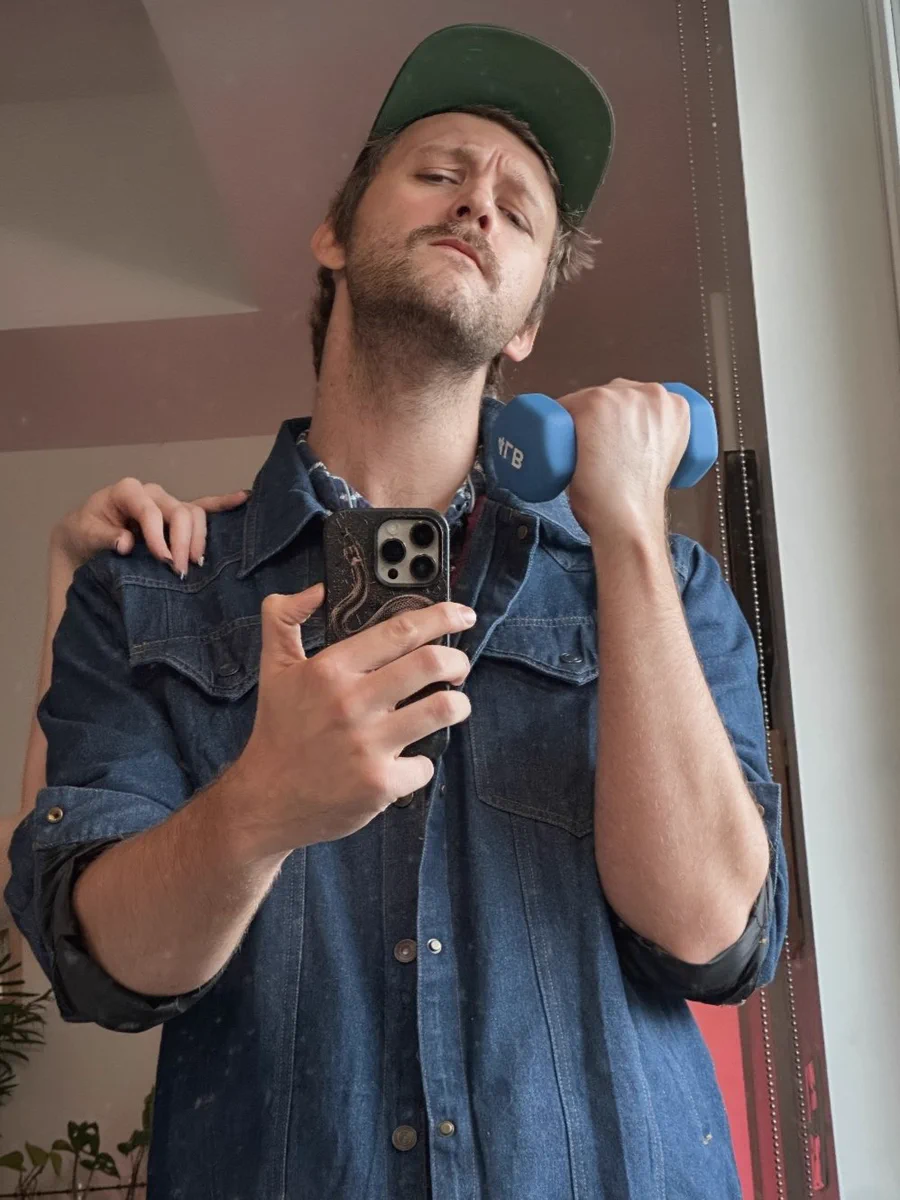 A fearless thought-leader of the Dark Gothic Woke movement. A 6'5" ridiculously handsome blond white boy with a killer jawline and a fierce look on his face. In his hand he holds a massive weight with ease, his bulging biceps nearly ripping through his jeans jacket. On his right shoulder, the hand of his beautiful pornstar wife gently rests (a feeling none of his haters will ever know). He looks at the camera with a defiant, confident gaze that says, "Go ahead, make my day, pal." It is clear he could destroy any conservative in the free marketplace of ideas with ease.