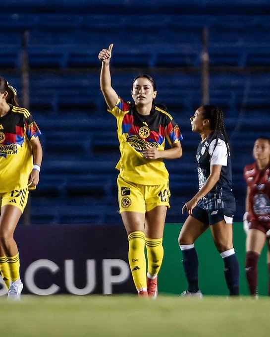 Scarlett con una camiseta de fútbol amarilla y negra del América Femenil con el número 14, levantando un brazo con el dedo señalador. Otros jugadores con camisetas amarillas y negras similares y uniformes del equipo contrario en colores oscuros son visibles en un campo de hierba. Los asientos azules del estadio están al fondo.