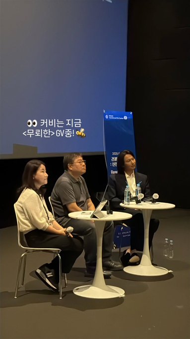 Three individuals on a stage with a large blue screen displaying text in Korean, including "12:45 무뢰한 무대인사" and "커뮤니티비프 18 COMMUNITY BIFF SEPTEMBER 2." One person holds a microphone, another sits at a table with water bottles and a laptop, and the third stands nearby. The setting includes a banner with the United Nations logo and text in Korean.