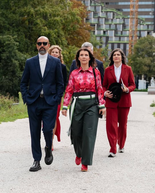 Esther Ouwehand standing at a podium, wearing a shirt with green, red, white, and black colors resembling the Palestinian flag. A microphone is positioned in front of her, and a stone wall is in the background. Esther Ouwehand walking outdoors with others, wearing a pink shirt with green and white accents and green pants, alongside individuals in suits and red outfits.