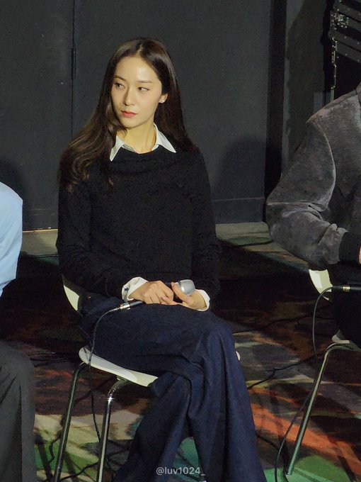 Jung Soojung sitting on a white chair, wearing a black sweater and dark blue pants, holding a microphone. Her long, straight hair is visible, and she appears to be at an event. A watermark "@luv1024_" is present in the bottom right corner.