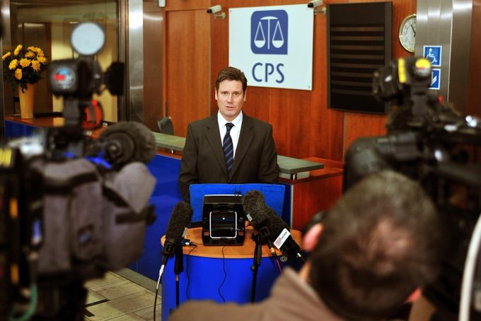 Keir Starmer sedí u stolu s mikrofony, na sobě má oblek a kravatu. Za ním je cedule s nápisem "CPS" a symbolem měřítka. V popředí jsou vidět kamery a mediální technika.