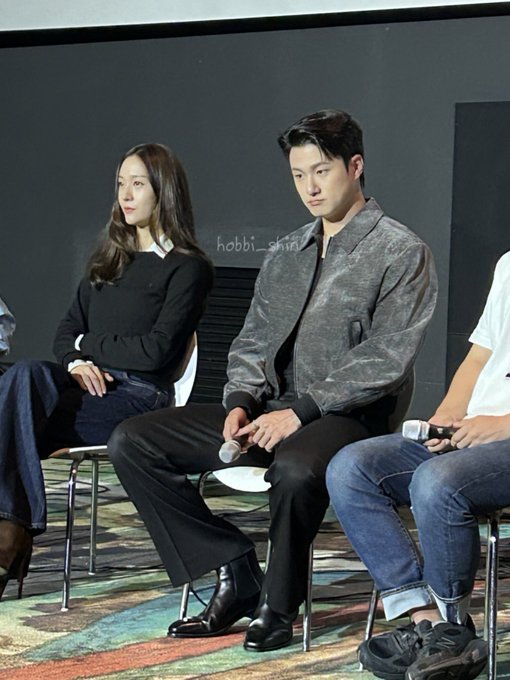 Shin Seung-ho sitting on a chair, wearing a shiny gray jacket, black pants, and black shoes, holding a microphone. A woman with long hair in a black top and blue pants sits to his left. A man in a white shirt and jeans sits to his right, holding a microphone. A watermark reading "hobbi_shin" is visible in the upper left corner.