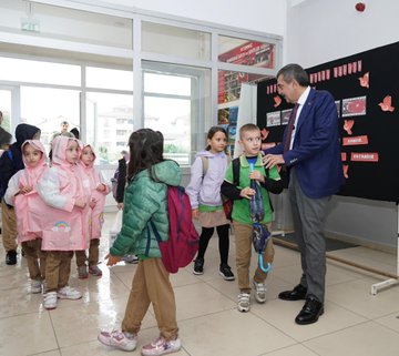 Children in colorful raincoats and backpacks walking outside a school building with a sign reading TOBB İlkokulu. A man in a blue suit interacts with students holding umbrellas indoors near a blackboard with red decorations. People in white coats and casual attire sit around a table in a meeting room with purple curtains. A group of individuals, including some in white coats, stands on steps outside the school building with a sign reading TOBB İlkokulu.