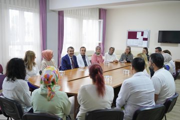 Children in colorful raincoats and backpacks walking outside a school building with a sign reading TOBB İlkokulu. A man in a blue suit interacts with students holding umbrellas indoors near a blackboard with red decorations. People in white coats and casual attire sit around a table in a meeting room with purple curtains. A group of individuals, including some in white coats, stands on steps outside the school building with a sign reading TOBB İlkokulu.
