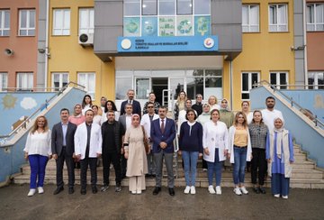 Children in colorful raincoats and backpacks walking outside a school building with a sign reading TOBB İlkokulu. A man in a blue suit interacts with students holding umbrellas indoors near a blackboard with red decorations. People in white coats and casual attire sit around a table in a meeting room with purple curtains. A group of individuals, including some in white coats, stands on steps outside the school building with a sign reading TOBB İlkokulu.