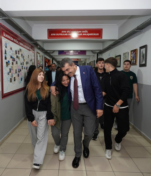 A man in a blue suit and red tie walking down a school hallway, shaking hands with a woman in a white coat. Students in casual clothing, some wearing jackets, walk alongside. A group of people, including individuals in white coats and suits, sit around a table in a room with lockers. A group of people, including men in suits and women in white coats, stand in front of a building with a sign reading "15 Temmuz Şehitler Anadolu Lisesi."