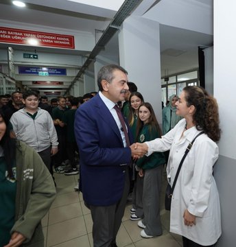 A man in a blue suit and red tie walking down a school hallway, shaking hands with a woman in a white coat. Students in casual clothing, some wearing jackets, walk alongside. A group of people, including individuals in white coats and suits, sit around a table in a room with lockers. A group of people, including men in suits and women in white coats, stand in front of a building with a sign reading "15 Temmuz Şehitler Anadolu Lisesi."