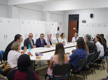 A man in a blue suit and red tie walking down a school hallway, shaking hands with a woman in a white coat. Students in casual clothing, some wearing jackets, walk alongside. A group of people, including individuals in white coats and suits, sit around a table in a room with lockers. A group of people, including men in suits and women in white coats, stand in front of a building with a sign reading "15 Temmuz Şehitler Anadolu Lisesi."