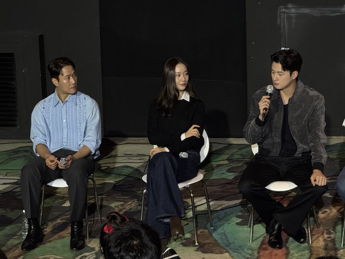 Three individuals sitting on chairs in a row on a patterned carpet. The person on the left wears a light blue shirt and dark pants, holding a small object. The person in the middle, a woman in a black sweater and dark pants, sits with legs crossed. The person on the right, in a dark jacket and pants, holds a microphone. Gang Tae-sik is implied to be present based on the post text.