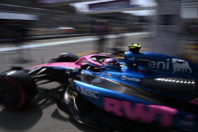A Formula One race car speeding on a track. The car is painted blue with pink accents, featuring visible logos including BWT and eni. The driver is visible inside the cockpit, wearing a helmet and racing suit.
