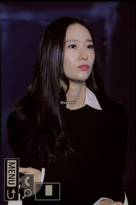 Jung Soojung with long dark hair, wearing a black outfit and white collared shirt. She has a thoughtful expression, touching her chin with one hand. A watermark with "@xxvxxl" is visible.