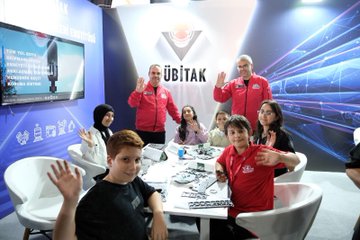 A girl wearing a VR headset and holding controllers, standing in a room with screens displaying virtual reality content. A boy interacting with a touchscreen display showing text and graphics. A group of people, including individuals in TÜBİTAK red jackets, sitting at a table with electronic components and screens. Two individuals holding cards with their photos and text. Blue and white decor with visible text on screens and walls.