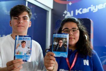 A girl wearing a VR headset and holding controllers, standing in a room with screens displaying virtual reality content. A boy interacting with a touchscreen display showing text and graphics. A group of people, including individuals in TÜBİTAK red jackets, sitting at a table with electronic components and screens. Two individuals holding cards with their photos and text. Blue and white decor with visible text on screens and walls.