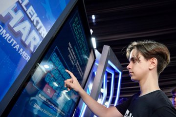 A girl wearing a VR headset and holding controllers, standing in a room with screens displaying virtual reality content. A boy interacting with a touchscreen display showing text and graphics. A group of people, including individuals in TÜBİTAK red jackets, sitting at a table with electronic components and screens. Two individuals holding cards with their photos and text. Blue and white decor with visible text on screens and walls.
