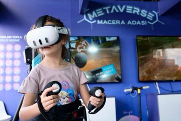 A girl wearing a VR headset and holding controllers, standing in a room with screens displaying virtual reality content. A boy interacting with a touchscreen display showing text and graphics. A group of people, including individuals in TÜBİTAK red jackets, sitting at a table with electronic components and screens. Two individuals holding cards with their photos and text. Blue and white decor with visible text on screens and walls.