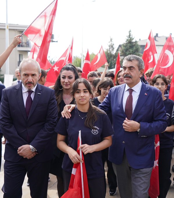 A group of people in formal attire standing together, cutting a ribbon at a school opening ceremony. Turkish flags and red-and-white balloons decorate the indoor space. A large banner displays "Esin Olcay Anadolu Lisesi Açılış Töreni" and images of individuals. Another image shows people, including children holding Turkish flags, walking outdoors with red flags in the background. A third image depicts a group of people posing on stairs inside a modern building with Turkish flags visible.