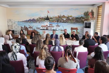 A group of students in yellow uniforms and teachers in formal attire stand in front of Mehmet Akif Ersoy Ortaokulu, a school with a blue and green building. Students reach out enthusiastically toward a man in a blue suit and red tie, Yusuf Tekin, in an outdoor setting with trees and a clear sky. Inside, Yusuf Tekin and others sit at a table, engaging with students and teachers in a room with a mural of Istanbul. Another image shows a large group of students, teachers, and Yusuf Tekin posing together outside the school.