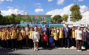 A group of students in yellow uniforms and teachers in formal attire stand in front of Mehmet Akif Ersoy Ortaokulu, a school with a blue and green building. Students reach out enthusiastically toward a man in a blue suit and red tie, Yusuf Tekin, in an outdoor setting with trees and a clear sky. Inside, Yusuf Tekin and others sit at a table, engaging with students and teachers in a room with a mural of Istanbul. Another image shows a large group of students, teachers, and Yusuf Tekin posing together outside the school.