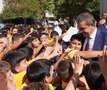 A group of students in yellow uniforms and teachers in formal attire stand in front of Mehmet Akif Ersoy Ortaokulu, a school with a blue and green building. Students reach out enthusiastically toward a man in a blue suit and red tie, Yusuf Tekin, in an outdoor setting with trees and a clear sky. Inside, Yusuf Tekin and others sit at a table, engaging with students and teachers in a room with a mural of Istanbul. Another image shows a large group of students, teachers, and Yusuf Tekin posing together outside the school.
