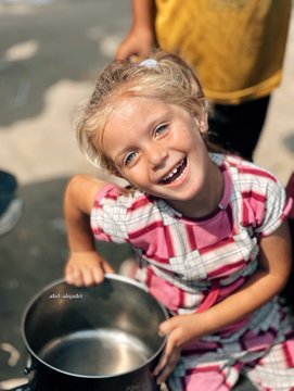 A young girl with blonde hair in pigtails, wearing a pink and white checkered dress, smiles brightly. She holds a large metal pot with both hands, sitting outdoors on a concrete surface. Other people, partially visible, wear colorful clothing, including a yellow shirt and a maroon robe, suggesting a communal setting. A watermark reading "abd_alqadri" is present on the image.