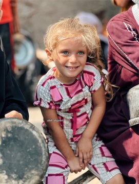 A young girl with blonde hair in pigtails, wearing a pink and white checkered dress, smiles brightly. She holds a large metal pot with both hands, sitting outdoors on a concrete surface. Other people, partially visible, wear colorful clothing, including a yellow shirt and a maroon robe, suggesting a communal setting. A watermark reading "abd_alqadri" is present on the image.