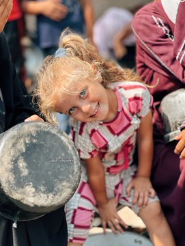 A young girl with blonde hair in pigtails, wearing a pink and white checkered dress, smiles brightly. She holds a large metal pot with both hands, sitting outdoors on a concrete surface. Other people, partially visible, wear colorful clothing, including a yellow shirt and a maroon robe, suggesting a communal setting. A watermark reading "abd_alqadri" is present on the image.