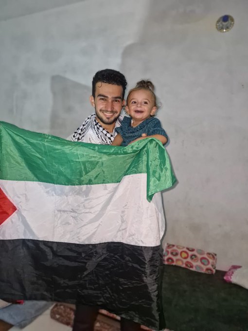 A man holding a child, both smiling. The man wears a checkered scarf and holds a large Palestinian flag with green, white, black, and red colors. A colorful pillow and a green mat are visible in the background.