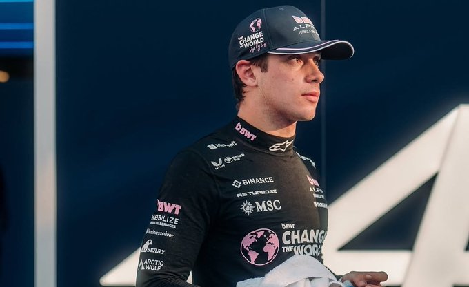Franco Colapinto wearing a black racing suit with multiple sponsor logos including BWT, Binance, and MSC. He has a dark cap with pink and white text and logos. The suit features a high collar and visible text such as "Change the World" on the chest.