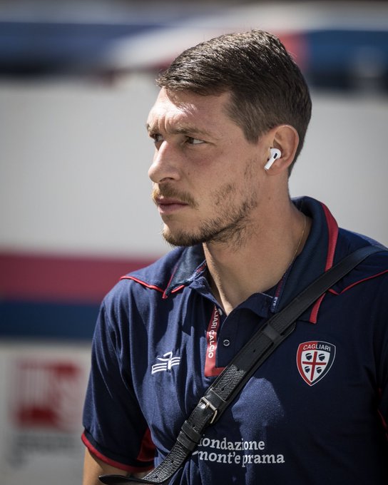 Andrea Belotti wearing a navy blue Cagliari jersey with red trim and a badge featuring a red and white shield. He has short brown hair, a light beard, and is wearing white earbuds. A black strap is visible across his shoulder.