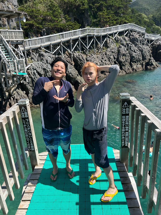 Chonmage Ramen Tabuchi and INI Matsuda Jin standing on a green wooden platform by the ocean. A wooden walkway extends over rocky cliffs and turquoise water. Signs with Japanese text are visible on the platform railing. People are swimming in the water in the background.