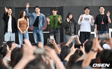 Six individuals standing on a stage, waving at a crowd. They are dressed in casual and stylish outfits, including a denim jacket, green shirt with tie, and leather jacket. The crowd in the foreground holds up phones, taking photos. A watermark reading "OSEN" is visible on the right side.