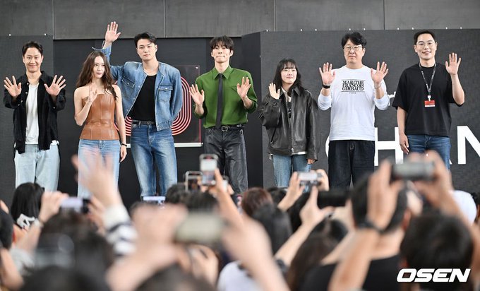 Six individuals standing on a stage, waving at a crowd. They are dressed in casual and stylish outfits, including a denim jacket, green shirt with tie, and leather jacket. The crowd in the foreground holds up phones, taking photos. A watermark reading "OSEN" is visible on the right side.