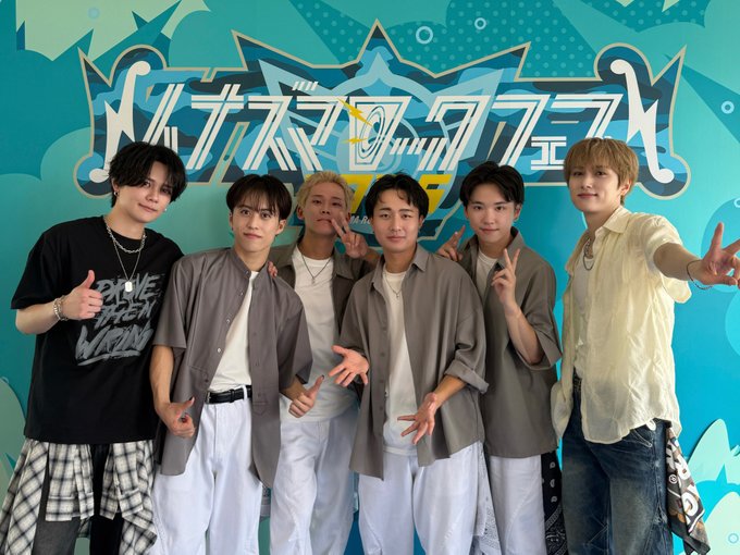 Seven individuals standing in front of a colorful blue background with Japanese text and graphics, including Takumi Ozaki, Kyosuke Fujimaki, and other members of INI. They wear casual outfits like black t-shirts, plaid shirts, and light-colored tops. The text on the background includes "イナズマロックフェス" and "IRF25".