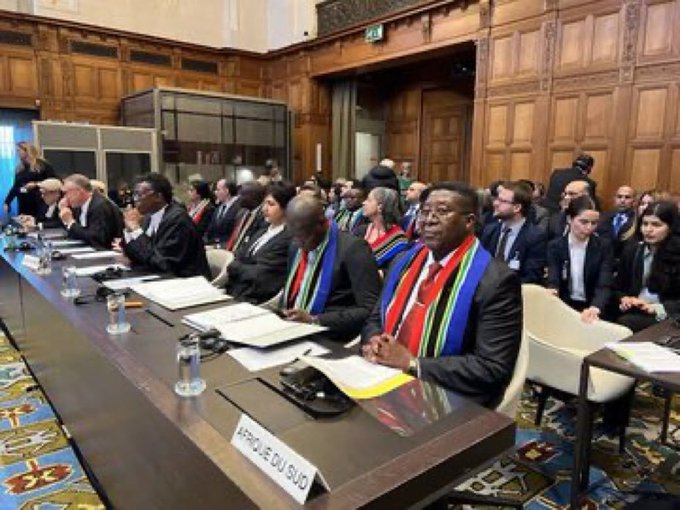 A formal meeting room with wooden paneling and a long table. Individuals in suits and some wearing sashes with red, green, blue, and yellow colors sit at the table. Nameplates, water bottles, and documents are on the table, with one visible nameplate reading "Afrique du Sud." A flag of Brazil with a green background, yellow diamond, blue globe, and white stars and text "Ordem e Progresso" is included.