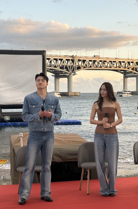 Shin Seung-ho and Jung So-jeong standing on a red stage by the water, holding microphones. They wear casual outfits, with Shin Seung-ho in a denim jacket and jeans, and Jung So-jeong in a brown top and jeans. A bridge and boats are visible in the background, along with a large screen and chairs on the stage.