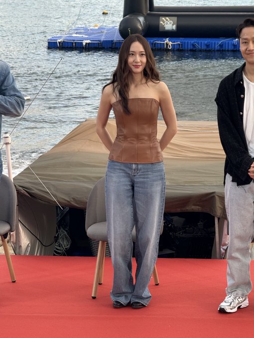 Song Hye-kyo standing on a red carpet, wearing a brown strapless top and loose blue jeans. She has long brown hair and black shoes. A body of water and a covered structure are visible in the background. Song Hye-kyo is also seated on a gray chair with wooden legs on the red carpet, dressed in the same outfit.