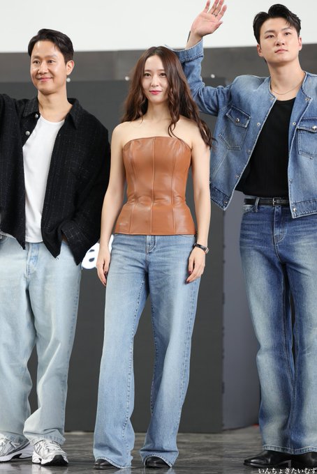Krystal Jung sitting on a white chair at a round table, wearing a brown strapless top and blue jeans, with a water bottle and mug on the table. Her long brown hair is styled loosely. In another image, Krystal Jung stands between two men in casual attire, all wearing blue jeans, with her raising one hand. A third image shows Krystal Jung seated, making a playful pout, with the same outfit and table setup. Text in the background reads "Fest 2024".