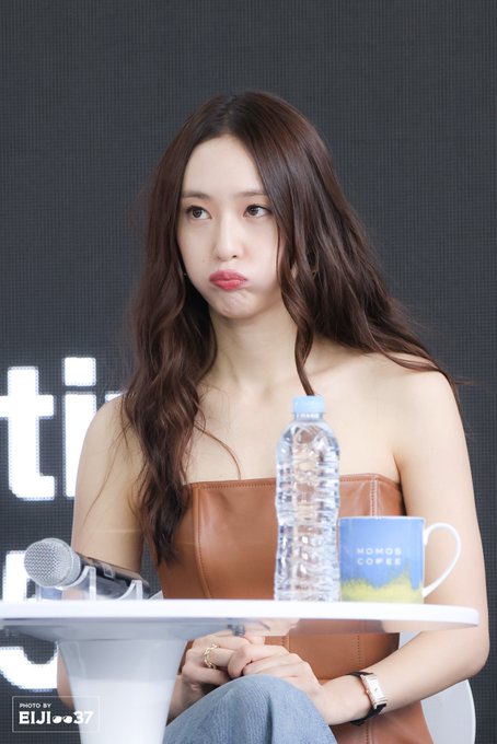 Krystal Jung sitting at a table during an event. She has long brown wavy hair and wears a strapless brown top and jeans. A microphone, a water bottle, and a mug with text are on the table. A watermark "EJI_0037" is visible in the bottom left corner.