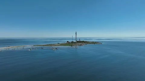 A small island, Vaindloo Island, surrounded by calm blue water in the Baltic Sea. The island has a narrow sandy shoreline and a central area with structures, including two tall towers or masts.