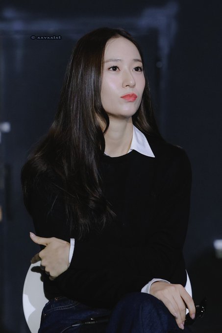 Jung Soojung sitting on a chair, wearing a black sweater over a white collared shirt and dark pants. Her long, straight dark hair falls over her shoulders. She has a thoughtful expression, with one hand touching her chin and the other resting on her lap. A watermark reading "@xxvxxlxt" is visible in the top left corner.