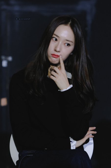 Jung Soojung sitting on a chair, wearing a black sweater over a white collared shirt and dark pants. Her long, straight dark hair falls over her shoulders. She has a thoughtful expression, with one hand touching her chin and the other resting on her lap. A watermark reading "@xxvxxlxt" is visible in the top left corner.