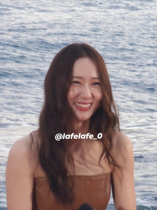 Jung Soojung with long brown hair, wearing a strapless brown top, smiling broadly. The background shows a calm ocean with blue water.