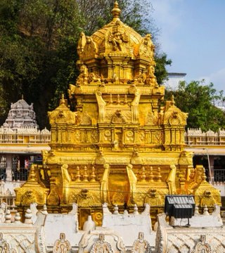 A golden temple gopuram adorned with intricate carvings and statues, featuring multiple tiers and a pointed top. The structure is surrounded by white stone railings and other temple buildings, with trees and hills visible in the background.