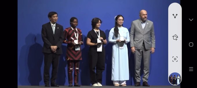 A certificate with the text "54th International Letter-Writing Competition for Young People" and "Reyyan Demiriz" as the first-place winner, featuring the United Nations logo. A gold medal and a black case with the United Nations emblem are beside the certificate. Four people, including Reyyan Demiriz, sit on a stage with Turkish and Vietnamese flags in the background. A large screen displays Reyyan Demiriz’s image with text about her first-place win and the UPU Congress, with an audience seated in front. A group of five people, including Reyyan Demiriz, stands on a stage with a blue backdrop.