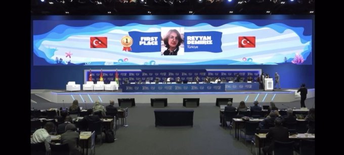 A certificate with the text "54th International Letter-Writing Competition for Young People" and "Reyyan Demiriz" as the first-place winner, featuring the United Nations logo. A gold medal and a black case with the United Nations emblem are beside the certificate. Four people, including Reyyan Demiriz, sit on a stage with Turkish and Vietnamese flags in the background. A large screen displays Reyyan Demiriz’s image with text about her first-place win and the UPU Congress, with an audience seated in front. A group of five people, including Reyyan Demiriz, stands on a stage with a blue backdrop.