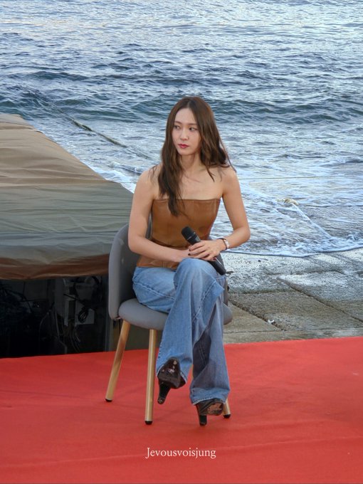 Jung Soojung sitting on a chair holding a microphone. She wears a strapless brown top, blue jeans, and black heels. The setting includes a red carpet, ocean waves, and a concrete pier.